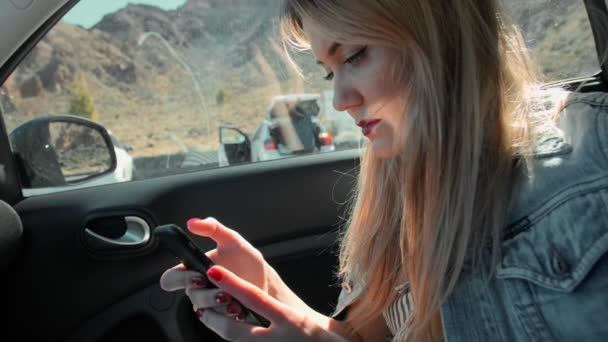 Mujer rubia atractiva en una chaqueta de mezclilla utiliza un teléfono inteligente mientras está sentado en el asiento del pasajero de un coche — Vídeo de stock