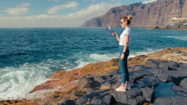 Mujer blogger rubia influencer dispara el surf en el video en un teléfono inteligente usando estabilizador de gimbal. En la orilla volcánica del océano. El concepto de hacer contenidos para las redes sociales — Vídeos de Stock