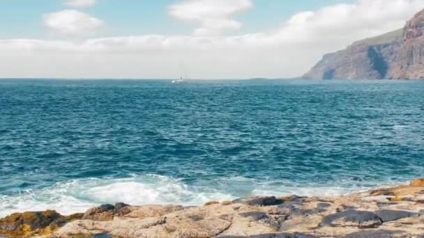 Lone yacht sta navigando oceano, roccia gigantesca sullo sfondo. Rocce gialle che fanno bollire il surf turchese. Il sole accecante di mezzogiorno illumina la riva rocciosa lavica. Il concetto di turismo attivo, ecologico e fotografico — Video Stock