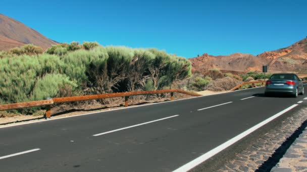 Auto non riconosciuta guida oltre la telecamera, cavalca lungo una strada asfaltata di montagna nel deserto. Sullo sfondo di una cima di un vulcano. Luminose indicazioni stradali. Sullo sfondo vulcanico — Video Stock