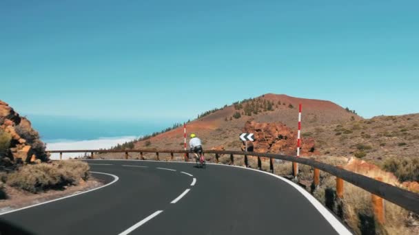 Atletizm sporcusu bir kır yolunda bisiklet sürüyor. Dönemece giriyor. Tanınmayan araba yaklaşıyor. Volkanik bir adanın manzarası, kayalık yol kenarı.. — Stok video