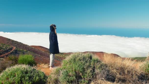 Ελκυστική γυναίκα στέκεται στην άκρη ενός γκρεμού. Η θέα πάνω από τα σύννεφα και το δάσος από ένα ψηλό βουνό. Πανόραμα θέα πάνω από τα σύννεφα και το δάσος από την κορυφή του βουνού. Κανάριοι Νήσοι — Αρχείο Βίντεο