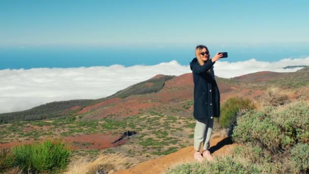 Attractive blonde woman makes selfie using a smartphone. Beautiful landscape with sea of clouds and the forest. Landscape with a sea of clouds above a valley and the forest covering a mountain ridge — Stock Video