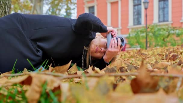 Beyaz, genç bir kadın fotoğrafçı eski model bir kameraya daha düşük bir açıdan fotoğraf çekiyor. Film ekipmanları üzerinde çekim yapmak dışında. Düşen yapraklarla sonbahar fotoğraf çekimi — Stok video