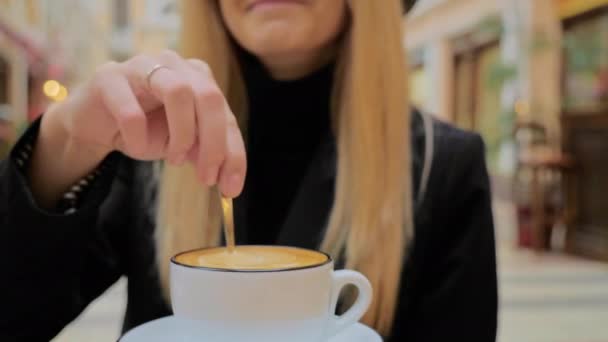 승인되지 않은 카푸치노 커피를 탄 세라믹 컵에 작은 숟가락으로 설탕을 휘젓는 백인 금발 여성 — 비디오