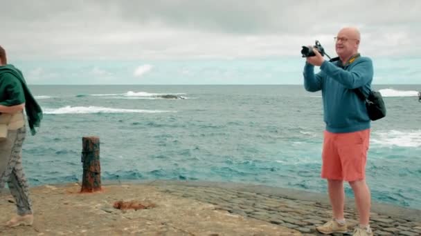Tenerife, Kanári-szigetek, Spanyolország - Január, 2019: Senor kopasz turista piros rövidnadrágban használja kamera az óceánon, A háttérben van egy gyanús ember, egy tolvaj. — Stock videók