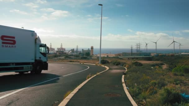 Tenerife, Kanári-szigetek, Spanyolország - 2019. január: Közúti teherszállítás. Szélmalmok elektromosság és olaj fúrótornyok a háttérben. Az energiakivonás fogalma — Stock videók
