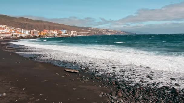 Sea surf. Ocean coast, waves on turquoise water and white foam. Drops fly into camera, volcanic black shore of stones and sand. coastal settlement from houses and hotels in far — Stock Video