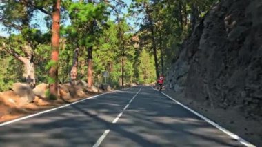 Tanınmayan bir bisikletçi dağ asfalt yolunda ilerliyor. Çerçevede hız sınırı var. Yol kenarındaki kayalık dağ tesisatları ve arka plandaki yeşil kozalaklı ağaçlar..