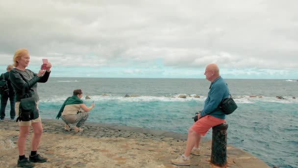 Teneriffa, Kanarieöarna, Spanien - januari 2019: Signora en äldre kvinna använder en smartphone för att få ett foto av en skallig man på havet. Flera turister tar bilder av sevärdheterna — Stockvideo