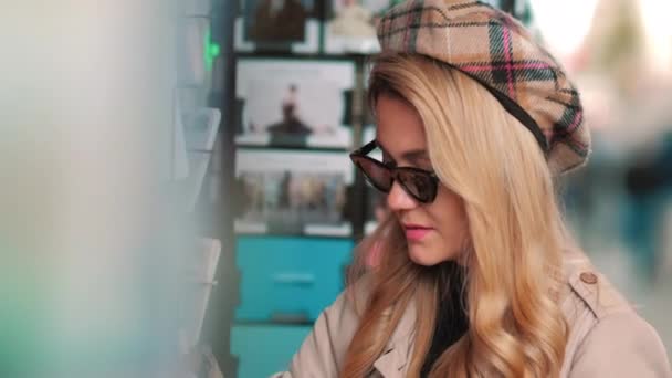Een jonge Parijzenaar kiest een ansichtkaart in een straatwinkel. Aantrekkelijk meisje in baret en zonnebril koopt een kaart ter herinnering aan de reis — Stockvideo
