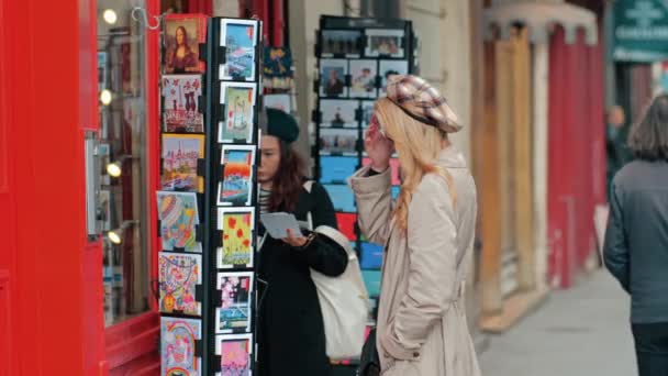 Paris, Fransa - Ekim 2019: İki çok ırklı kız sokakta küçük bir hediyelik eşya dükkanında kartpostal seçiyor. Asyalı ve Parisli stili bereler. — Stok video