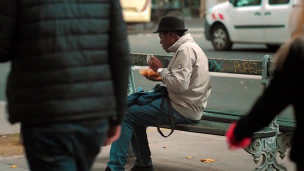 Párizs, Franciaország - Október, 2019: hajléktalan fekete férfi kalapban eszik ételt az utcán. Különböző emberek járnak erre. Luxus autó halad a háttérben. A menekültek és a migránsok fogalma. — Stock videók
