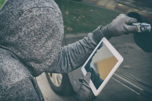 Вор в капюшоне пытается взломать систему безопасности машины с помощью планшета — стоковое фото