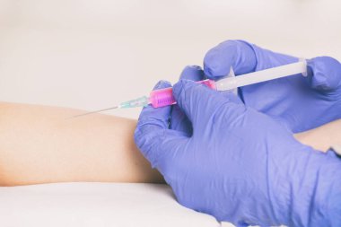 Doktorun elleri hastanın ilacıyla birlikte kullanıma hazır bir şırınga tutuyor. Hastanın eli arka planda görünüyor.