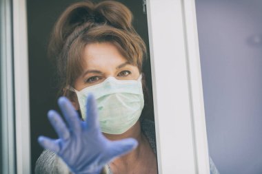 Korumalı yüz maskesi takan ve Corona virüsü salgını yüzünden evinden dışarı bakan bir kadın. Ev karantinası ve kendini izole etme kavramı
