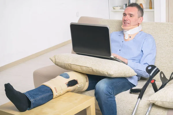 Man with leg in neck brace, knee cages and crutches for stabilization and support resting on a sofa with laptop.