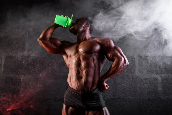 Siyah arka planda dumanlı vücut geliştirici. Afrika kökenli Amerikalı erkek sporcu bir sallamacıdan spor beslenmesi içiyor — Stok fotoğraf