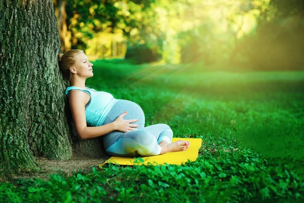 Молодая мама, беременная девушка, отдыхает в парке возле дерева, после занятий йогой — стоковое фото