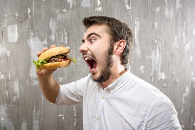 Sakallı genç adam gri arka planda ev yapımı hamburger yiyor.