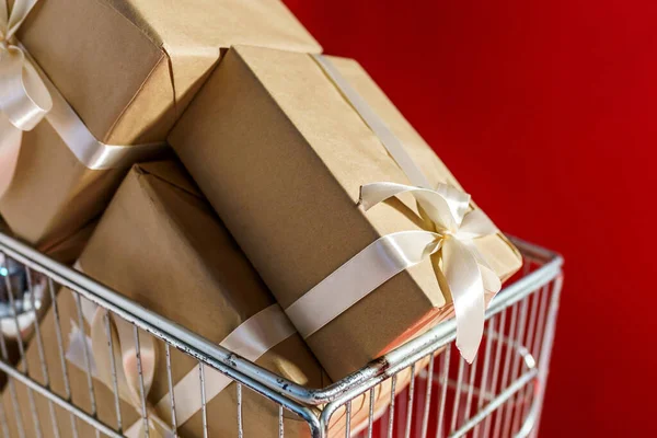 Dárky v řemeslném papíru s bílou páskou v supermarketu vozíku na červeném pozadí — Stock fotografie