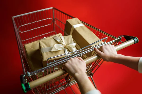 Girl holds hands a supermarket trolley with gifts packed in craft paper with a white ribbon on a red background — Stock Photo, Image