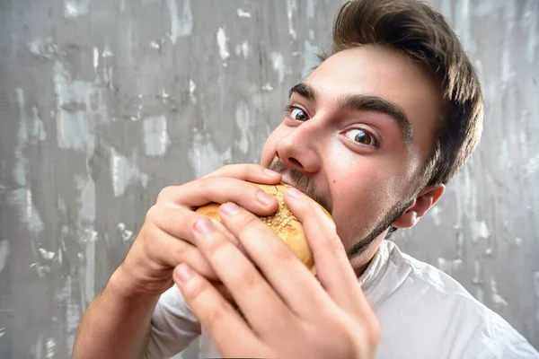 Молодий хлопець з бородою їсть домашній гамбургер на сірому фоні — стокове фото
