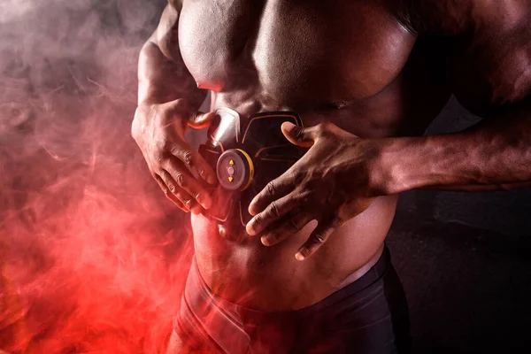 Bodybuilder αρσενικό Αφρικής χρησιμοποιεί ηλεκτρονική ζώνη μυϊκό διεγέρτη κοιλιακούς μυς σε μαύρο φόντο με κόκκινο καπνό — Φωτογραφία Αρχείου