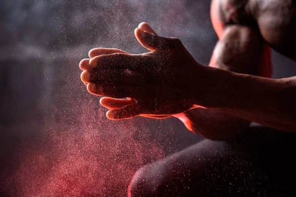 Bodybuilder African American uses hand magnesia. A man sits on a black background with red smoke — стокове фото