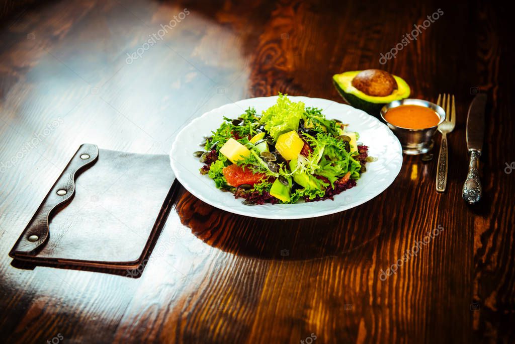Restaurant menu in a leather folder on a wooden table near a salad with fresh vegetables and fruits