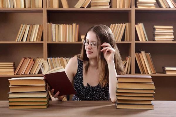 Молодая студентка читает учебник, сидя за столом с большим количеством книг в библиотеке — стоковое фото