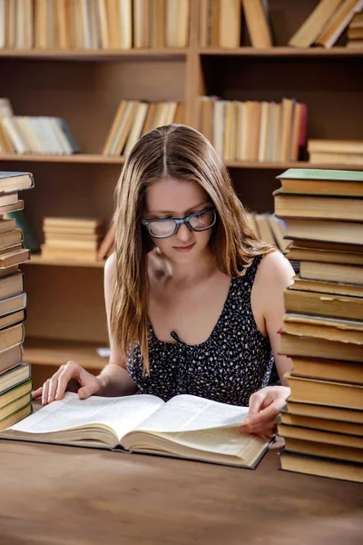 Joven estudiante lee un libro de texto sentado en una mesa con muchos libros en la biblioteca — Foto de Stock