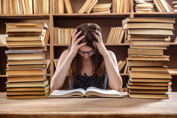 Молодая студентка читает учебник, сидя за столом с большим количеством книг в библиотеке
