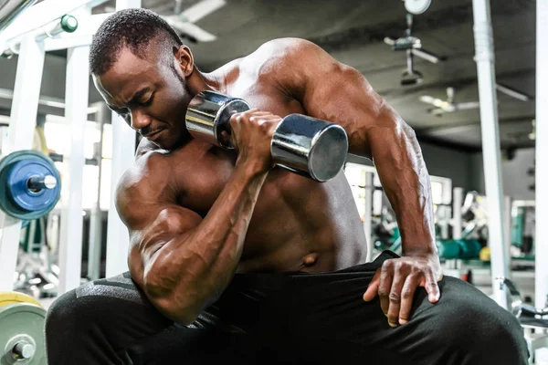 Spor salonundaki bir bankta otururken, Afrikalı erkek sporcu halterlerle egzersiz yapıyor. — Stok fotoğraf