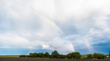 Ağaçların ve tarımsal alanların üzerinde iki gökkuşağı ve arka planda bulutlu gökyüzü