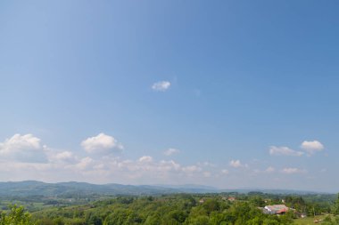 Tepe manzarası ağaç örtüsüyle doludur. Bazı evler ve bulutlu mavi gökyüzü vardır.