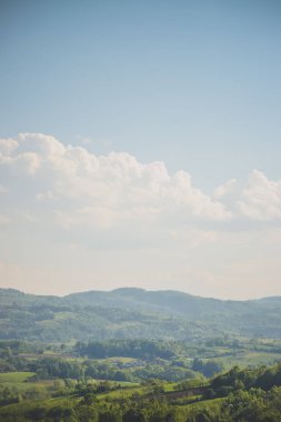 Tepe manzarası ağaç örtüsüyle doludur. Bazı evler ve bulutlu mavi gökyüzü vardır.