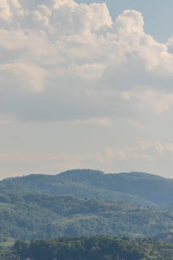 Tepe manzarası ağaç örtüsüyle doludur. Bazı evler ve bulutlu mavi gökyüzü vardır.