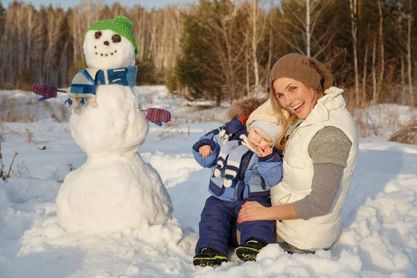 Happy mother and baby in winter park. family outdoors with snowman — Stock Photo, Image