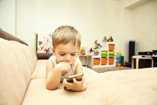 Un niño pequeño mirando al teléfono inteligente acostado en el sofá en casa . —  Fotos de Stock