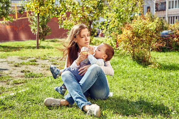 Ung mamma sitter på gräset och matar sitt barn från en flaska. Mamma och son i en park. — Stockfoto