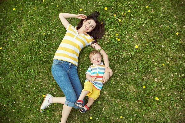 Utomhusporträtt av en mor med sitt barn. Mamma och son går i en sommarpark på gräset med gula maskrosor. ovanifrån. — Stockfoto