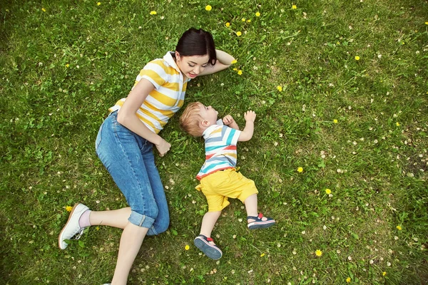 Utomhusporträtt av en mor med sitt barn. Mamma och son går i en sommarpark på gräset med gula maskrosor. ovanifrån. — Stockfoto