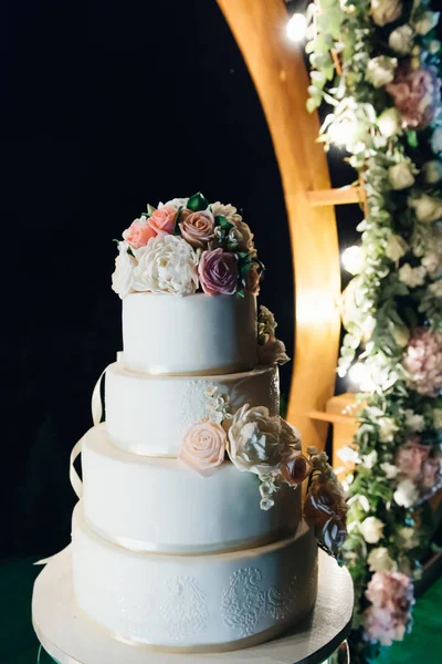 Torta blanca de lujo de cuatro niveles con flores en arco de boda de noche —  Fotos de Stock