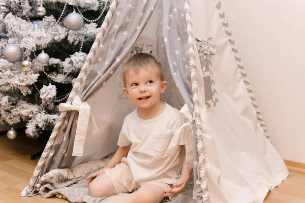 一个可爱的小男孩正坐在靠近圣诞树的一座帐篷小屋的儿童房里 — 图库照片