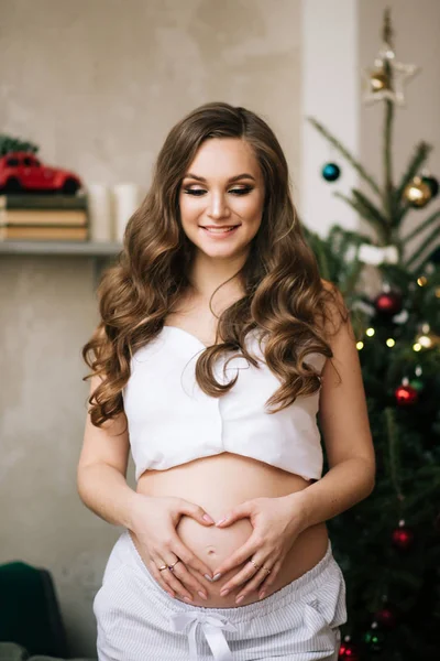 Güzel, genç, şık, beyaz pijamalı hamile bir kız Noel ağacıyla şık bir iç mekanda kanepede oturuyor.