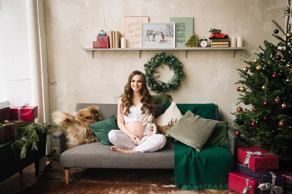 美しく若いです 白いパジャマでスタイリッシュな妊娠中の女の子はクリスマスツリーとスタイリッシュなインテリアでソファに座っています — ストック写真