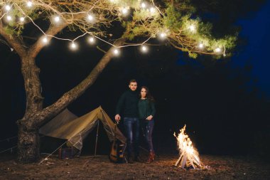 Aşık genç bir çift, ormanda bir çadırın önünde eski bir çelenkle kamp kuruyor. Çok gürültülü bir fotoğraf, seçici bir odak noktası var.