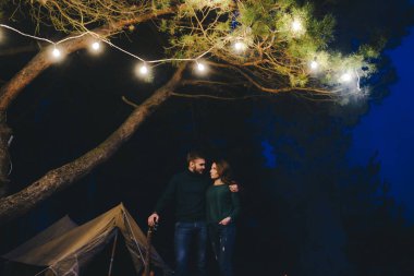 Aşık genç bir çift, ormanda bir çadırın önünde eski bir çelenkle kamp kuruyor. Çok gürültülü bir fotoğraf, seçici bir odak noktası var.