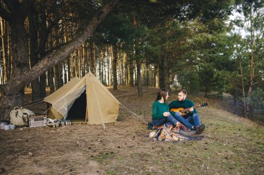 Aşık genç bir çift kamp ateşi başında çadır kurmuş gitar çalıyor.